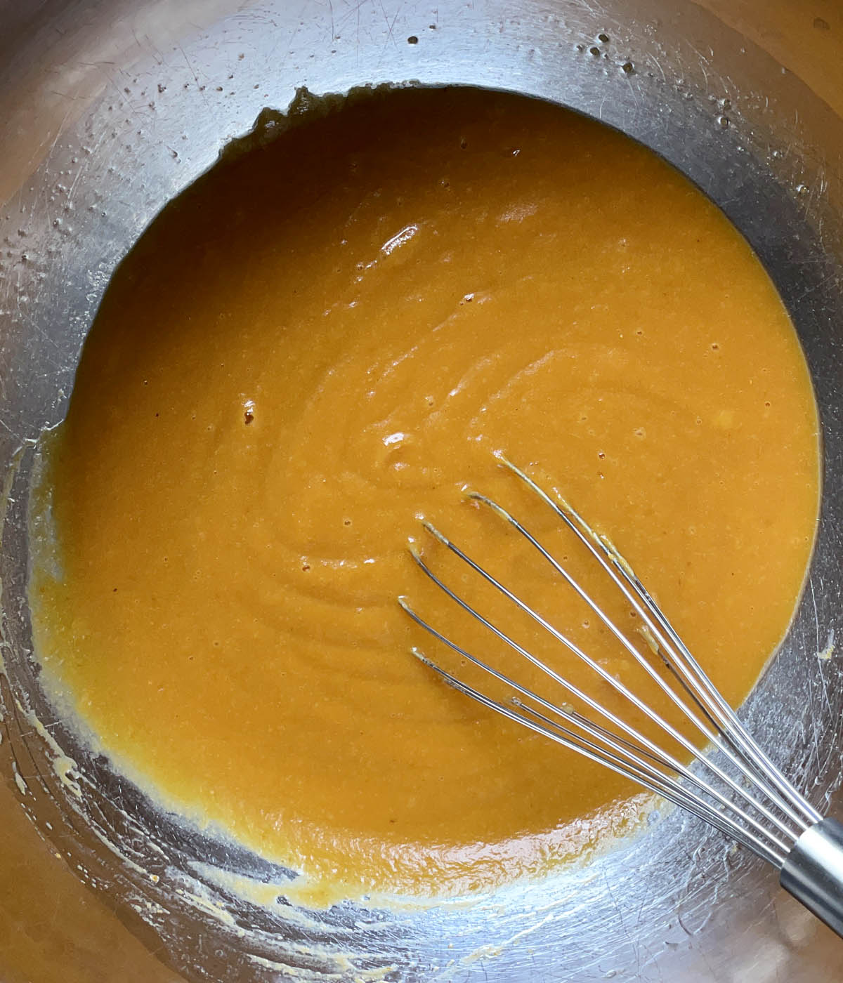 A metal round bowl containing an orange liquid and a whisk.