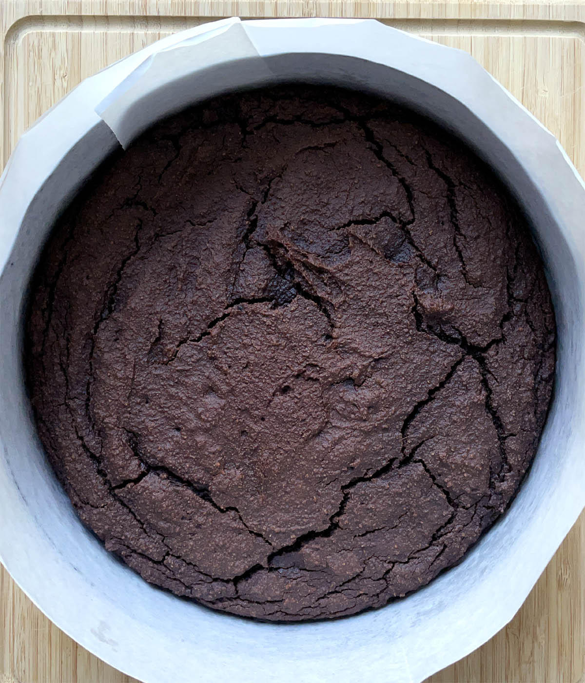 A brown baked chocolate cake in a round pan lined with white paper.