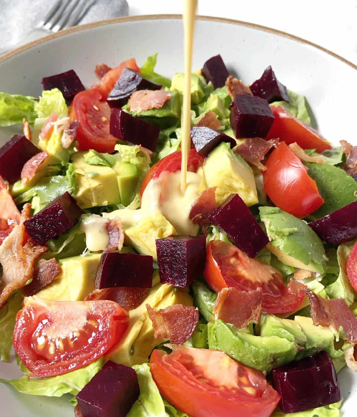 Yellow dressing being poured over a salad of red tomatoes, purple beets, green avocado chunks, and cooked bacon.