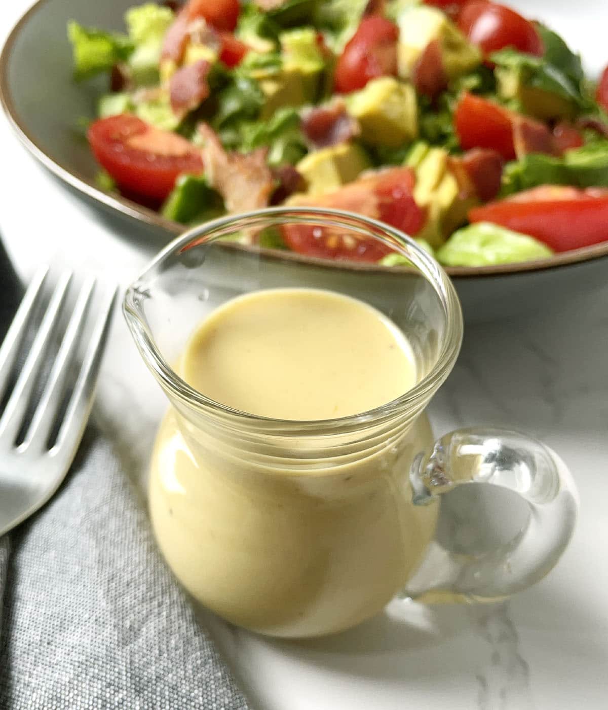 A glass pitcher containing yellow honey mustard dressing, next to a fork and a plate containing a salad.