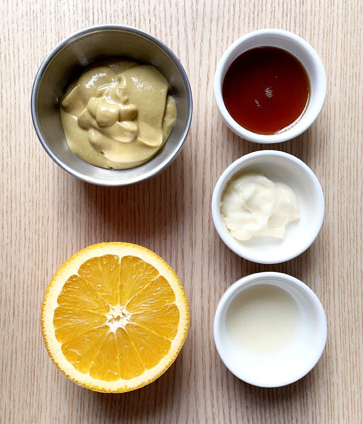 Half an orange, a metal bowl containing mustard, a white bowl containing brown honey, a white bowl containing white mayonnaise, a white bowl containing vinegar.