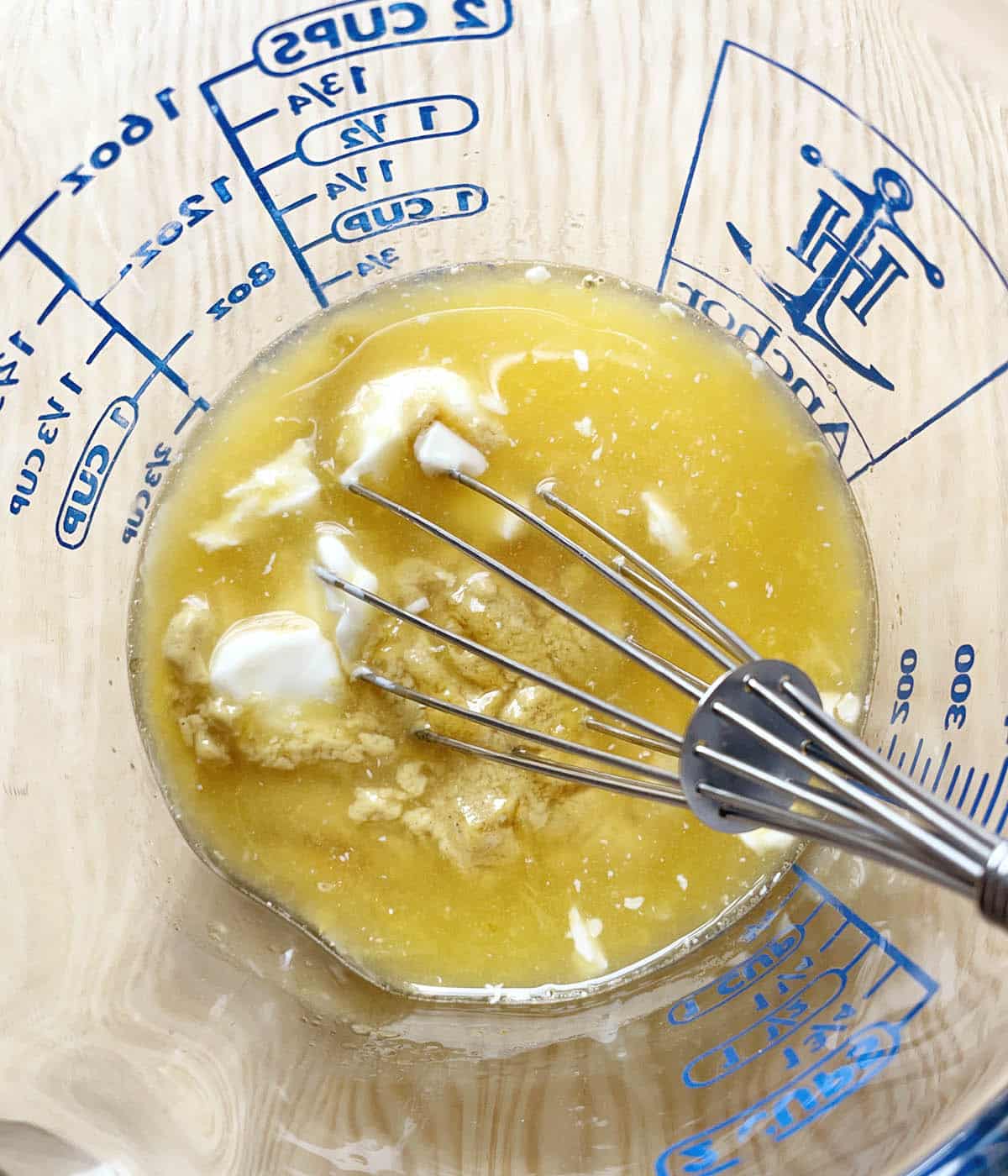 A glass measuring cup containing a metal whisk, orange juice, mayonnaise, and mustard.