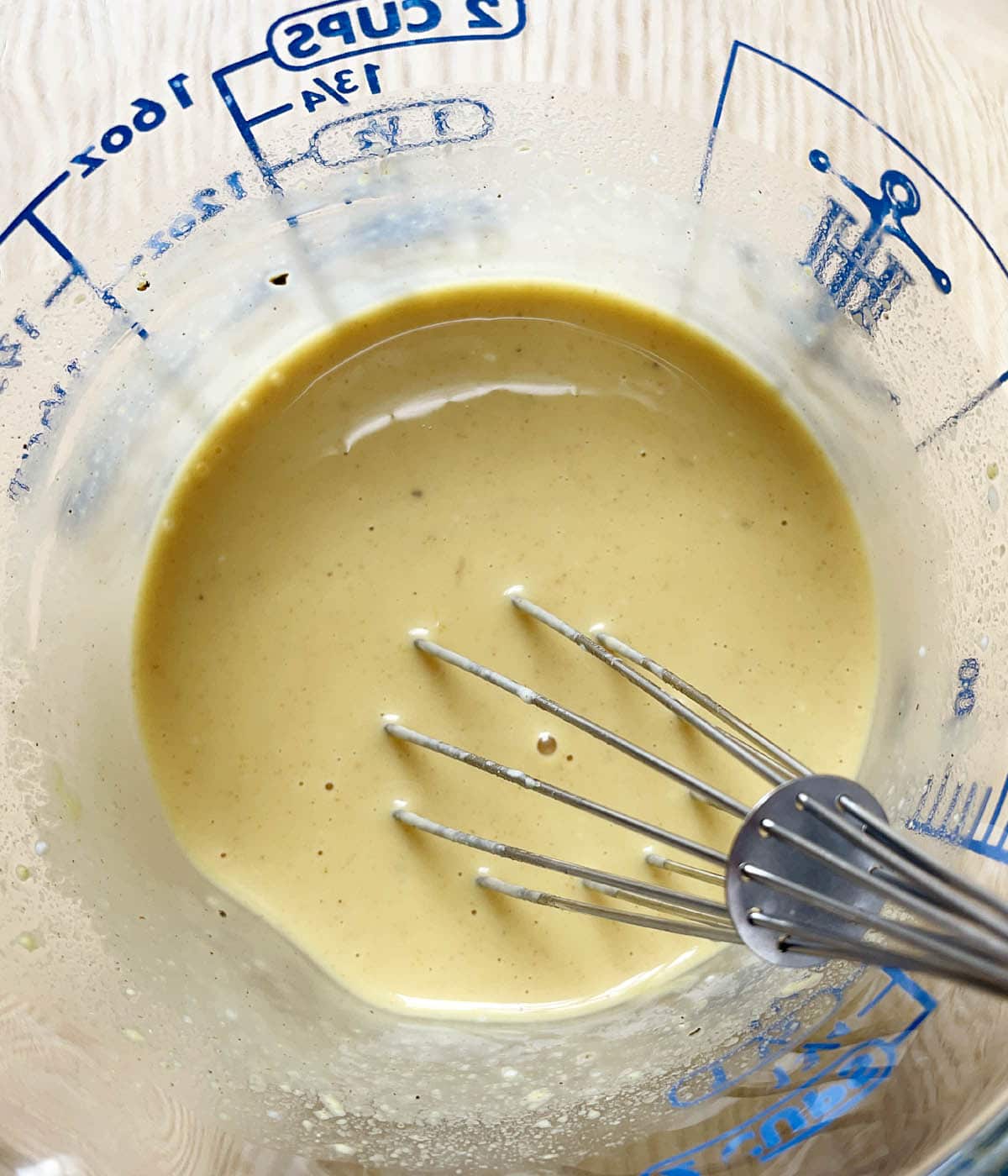 A glass measuring cup containing a whisk set in yellow honey mustard dressing.