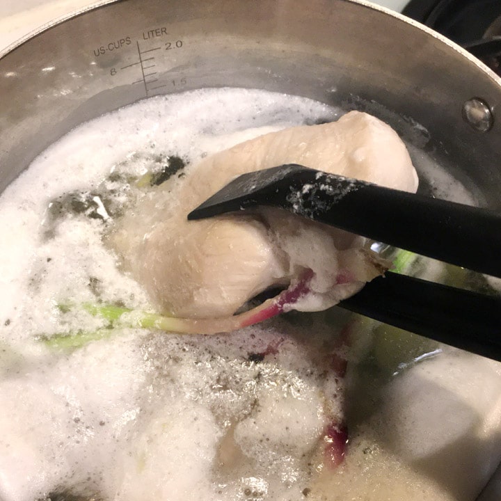 Black tongs turning chicken breast over in a pot containing foamy water