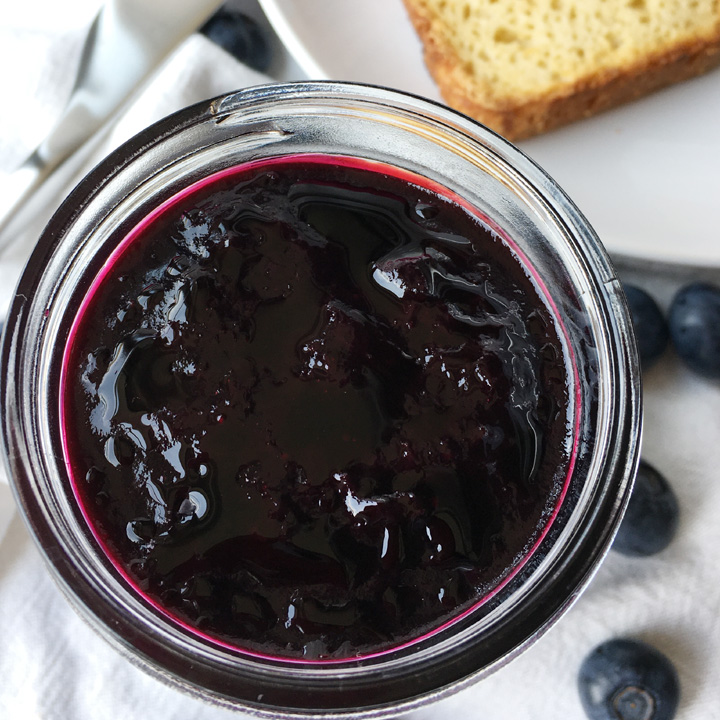 Looking down on an open jar of purple blue jam, next to toaste on a white plate