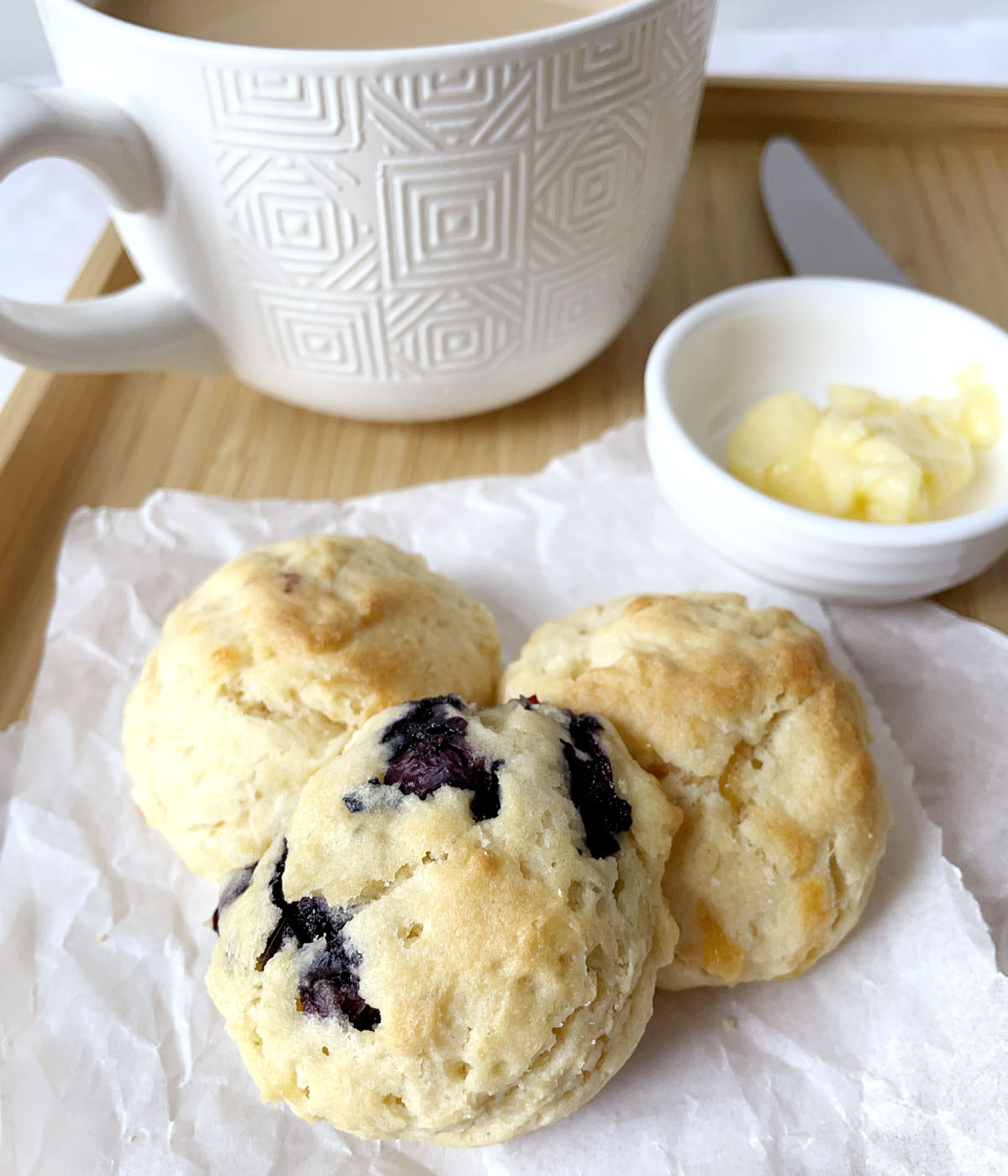 One blueberry scone and two cheese tea scones on white parchment paper, next to a dish of yellow butter and a white mug of tea.