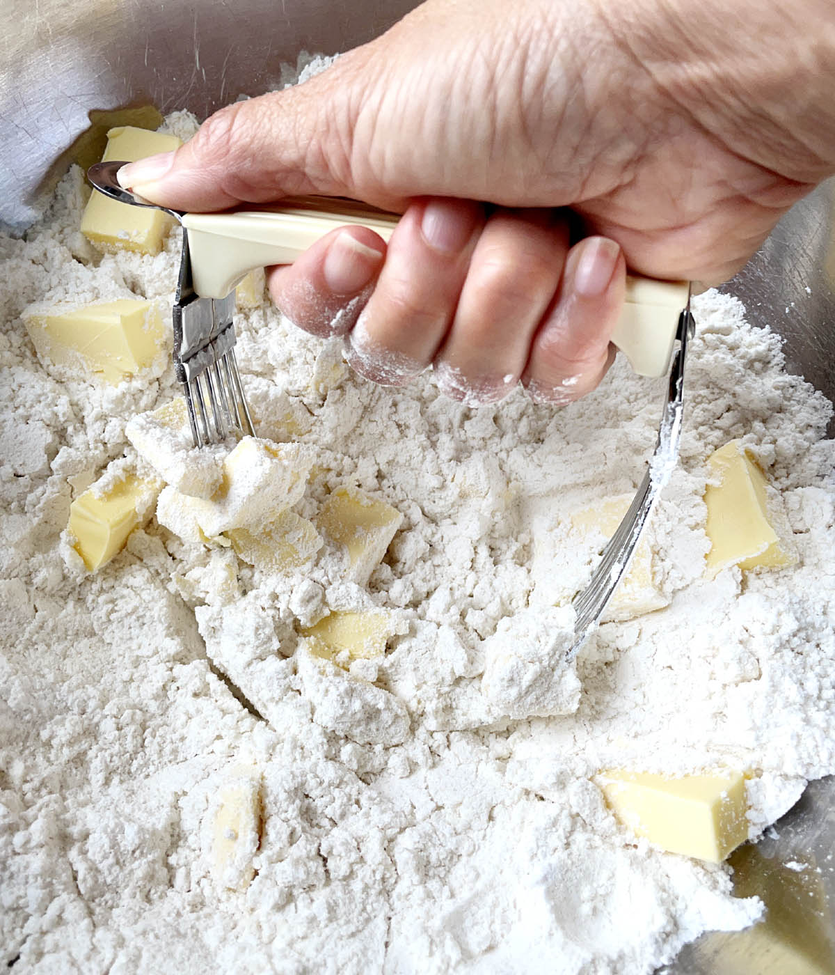 A hand holding a pastry cutter, cutting into butter in a flour mixture in a bowl.