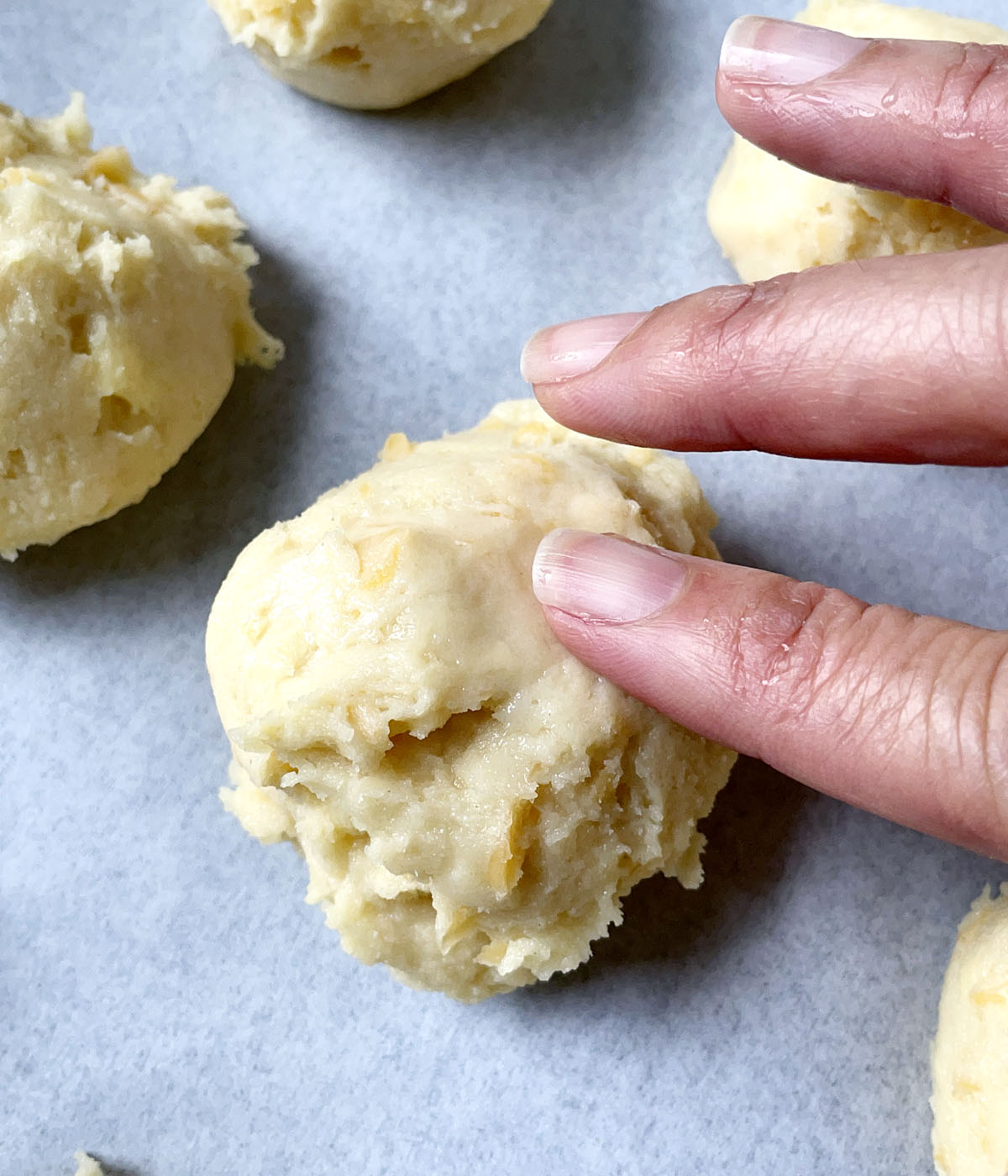 Fingers pressing down on the jagged edges of scoops of dough on white parchment paper.