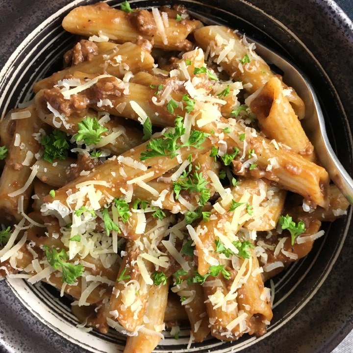 A dark round bowl containing beef and pasta, topped with grated white cheese and chopped green parsley