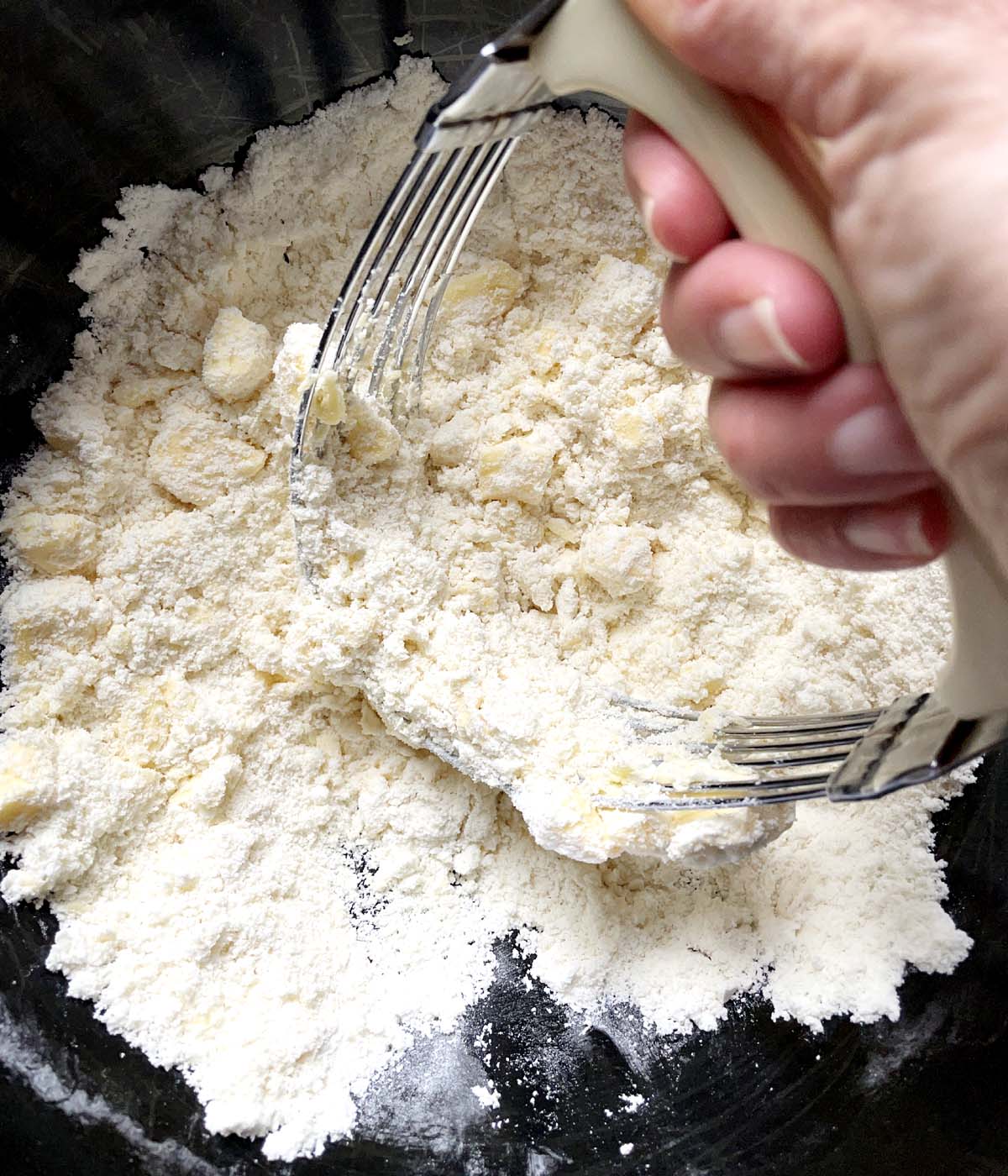 A hand holding a metal pastry blender cutting into butter and flour in a black bowl.
