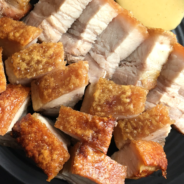 A black dish containing chunks of roast pork belly with orange crispy skin, next to some yellow mustard
