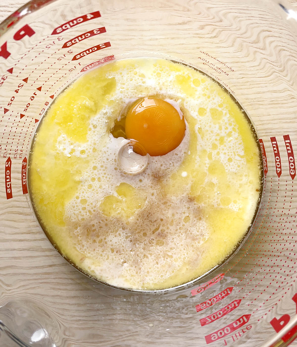 A round glass measuring cup containing an egg and yellow and white liquid.