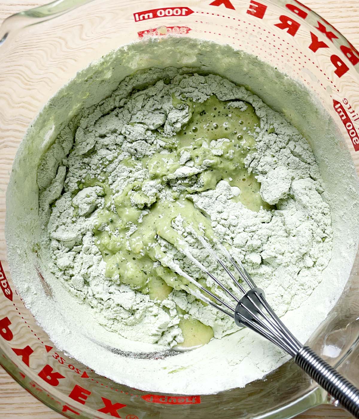 A whisk mixing white and green flour ingredients into a liquid in a glass measuring cup.