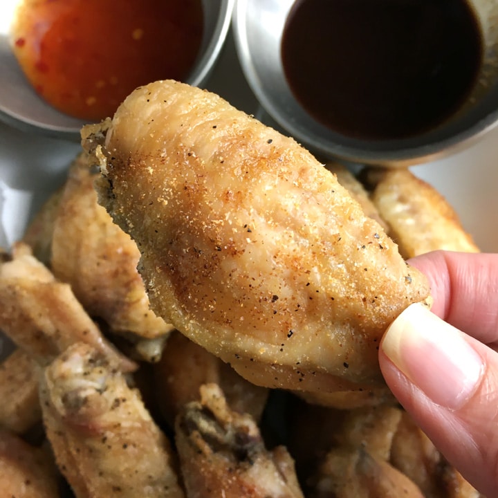 Close-up of fingers holding a brown baked chicken wing over a plate of chicken wings and two bowls with sauces
