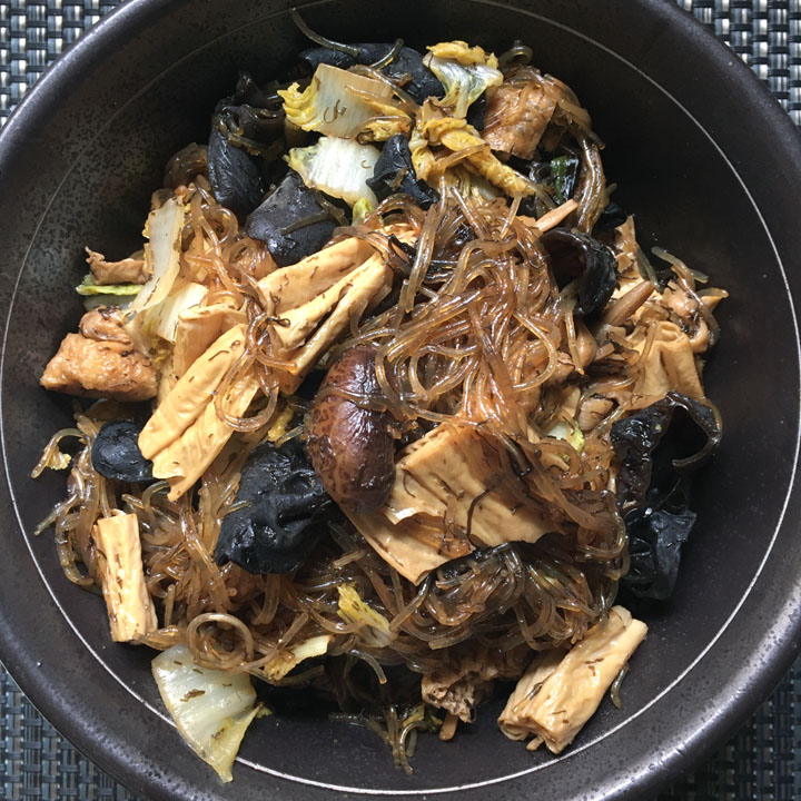 A dark round bowl containing noodles, mushrooms, chopped cabbage