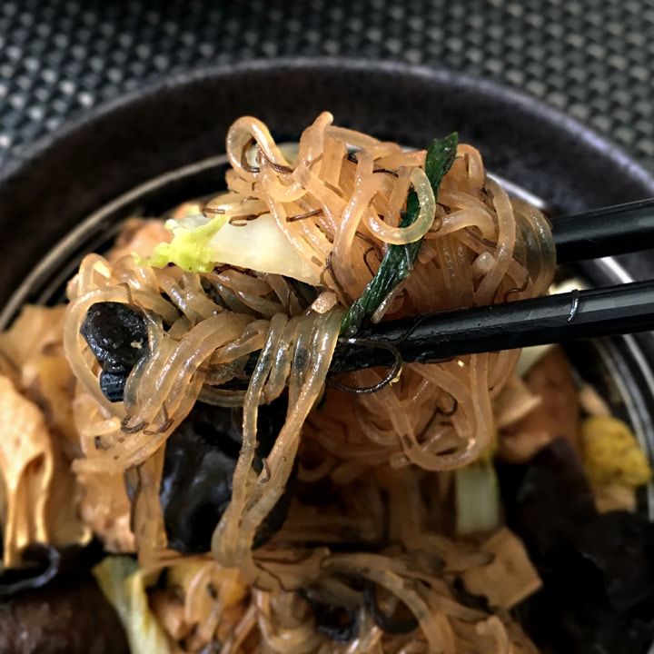 Close-up of a pair of black chopsticks holding brown noodles, chopped cabbage, green onion, black fungus