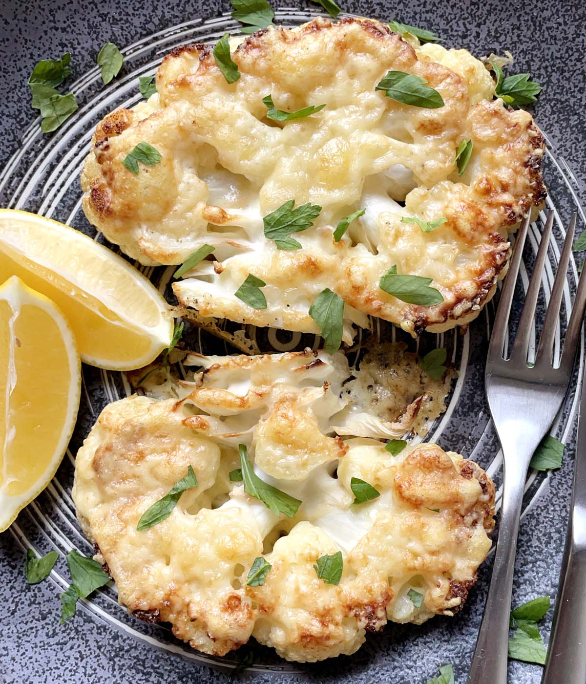 Two parmesan roasted cauliflower steaks, two lemon wedges, and a fork and knife on a dark round plate.
