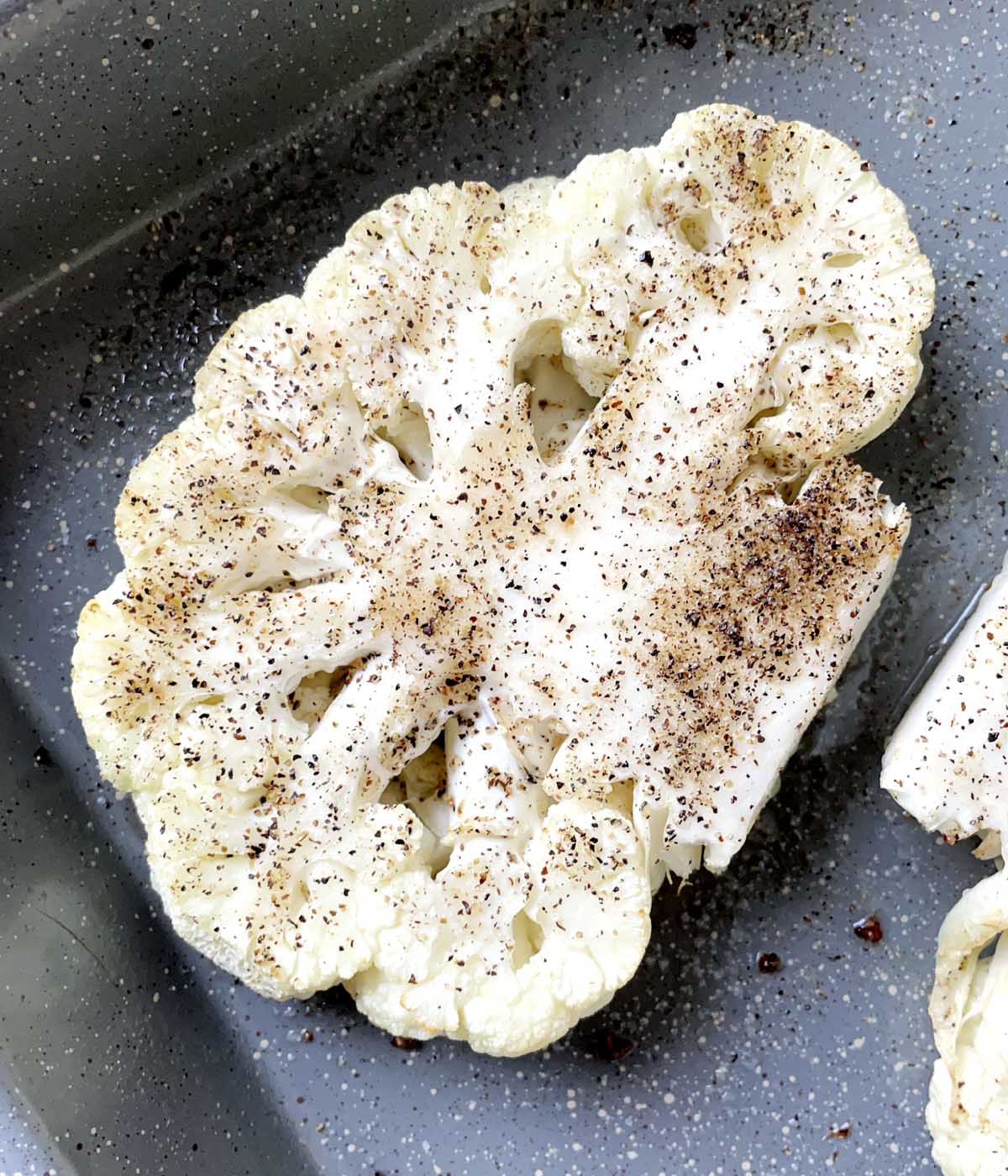 A roasted slice of cauliflower topped with back pepper in a grey roasting pan.