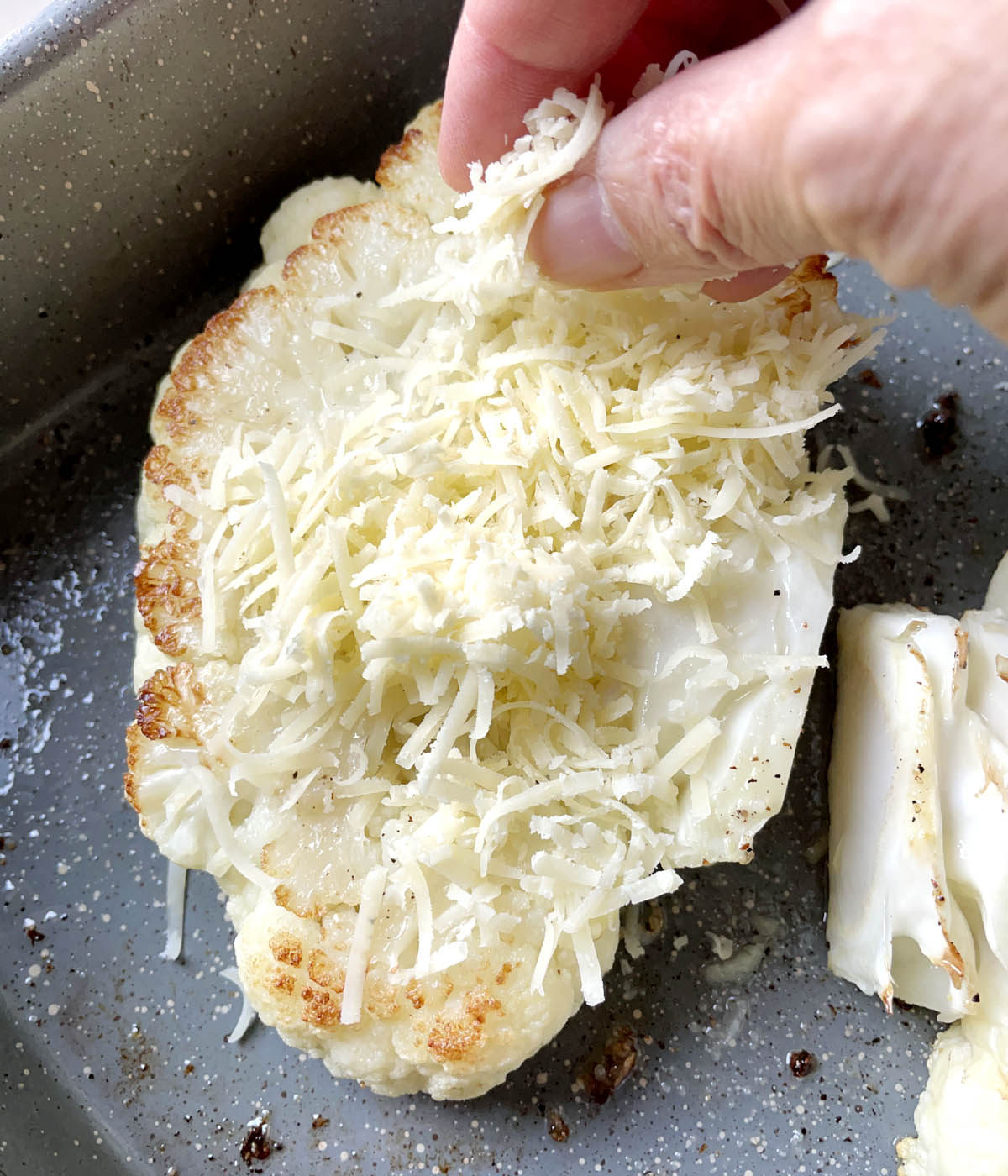 A hand spreading grated parmesan cheese over a cauliflower slice.
