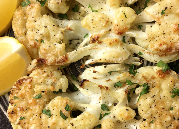 Close-up of two roasted cauliflower steaks on a round plate with yellow lemon wedges
