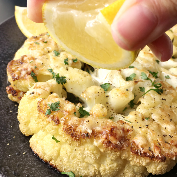 Close-up of a hand squeezing juice from a yellow lemon wedge over a roasted cauliflower steak on a dark plate
