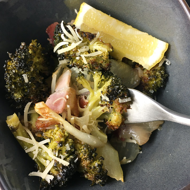 A grey bowl containing with a fork poking into roasted broccoli and bacon and onions, a yellow lemon wedge