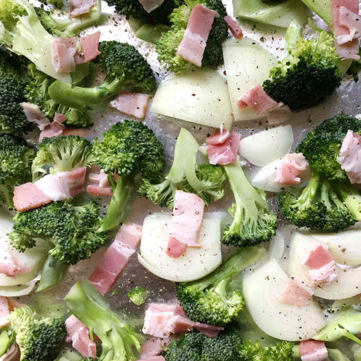 Raw green broccoli, raw bacon pieces, raw onion chunks on a foil-lined pan, sprinkled with salt and pepper