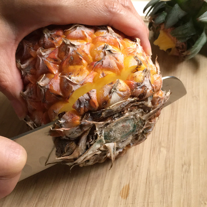 A hand holding a pineapple, a knife cutting the bottom off a pineapple