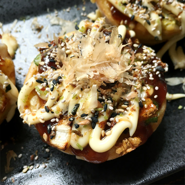 A muffin shaped okonomiyaki topped with brown sauce, white mayonnaise, sesame seeds, and brown flakes