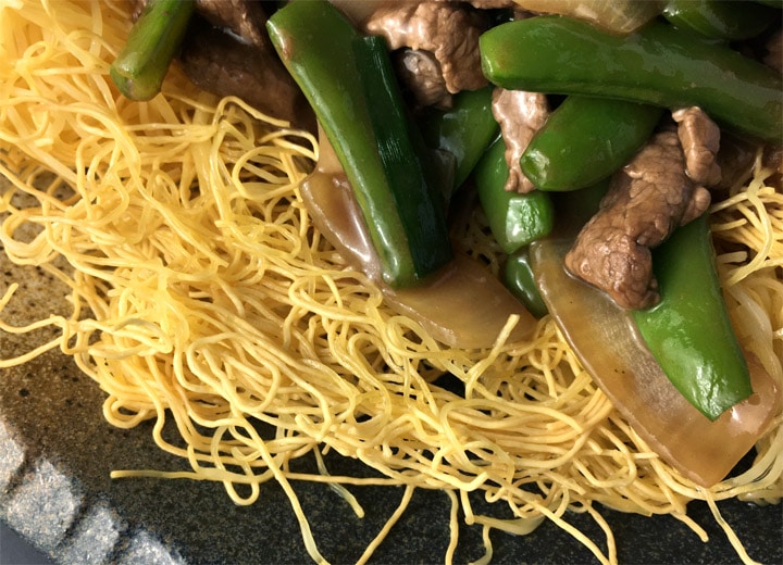 Close-up of yellow noodles topped with beef, green pea pods, and onions