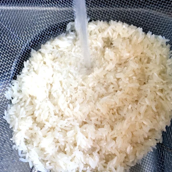 Uncooked rice being rinsed with water in a colander.