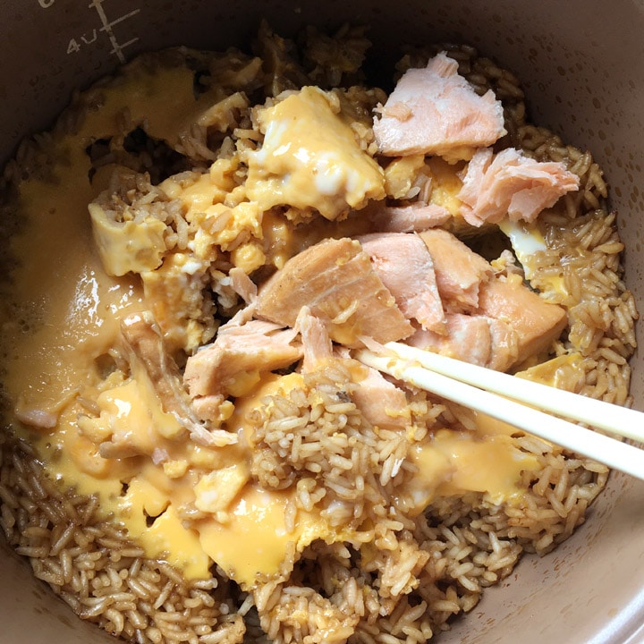 A pair of chopsticks in a pot with broken up salmon, yellow eggs, and rice.