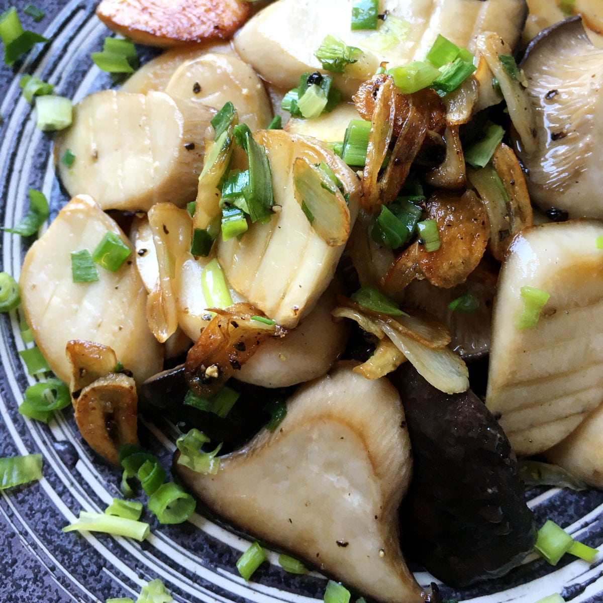 A round dark grey bowl containing cooked mushroom slices, fried garlic slices, and chopped green onions.