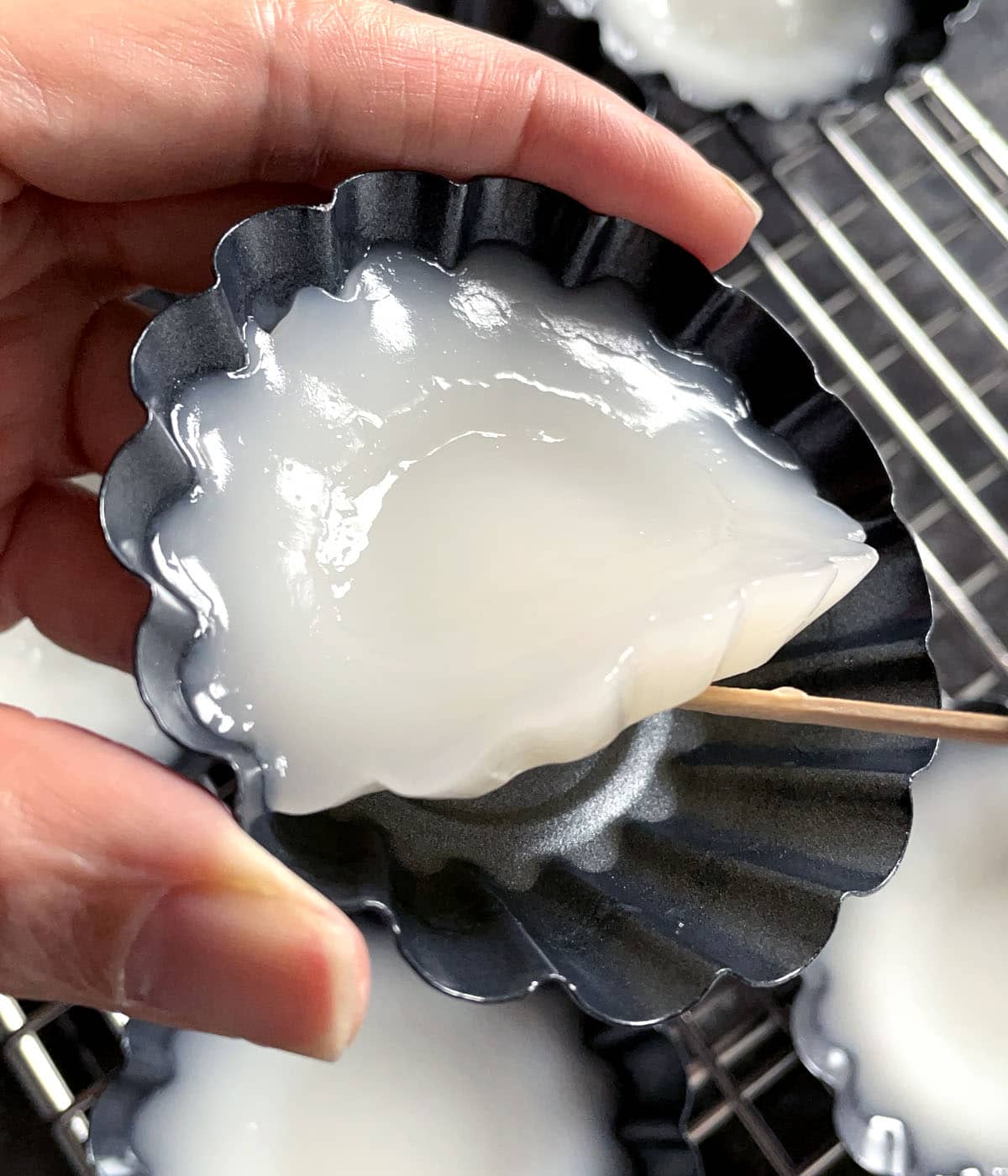 A toothpick loosening a round white jelly pudding from a dark dish.