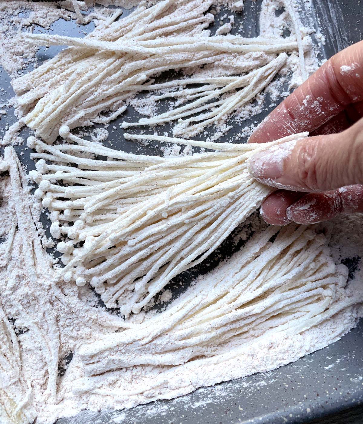 A hand holding a bundle of raw enoki mushrooms coated in white flour.