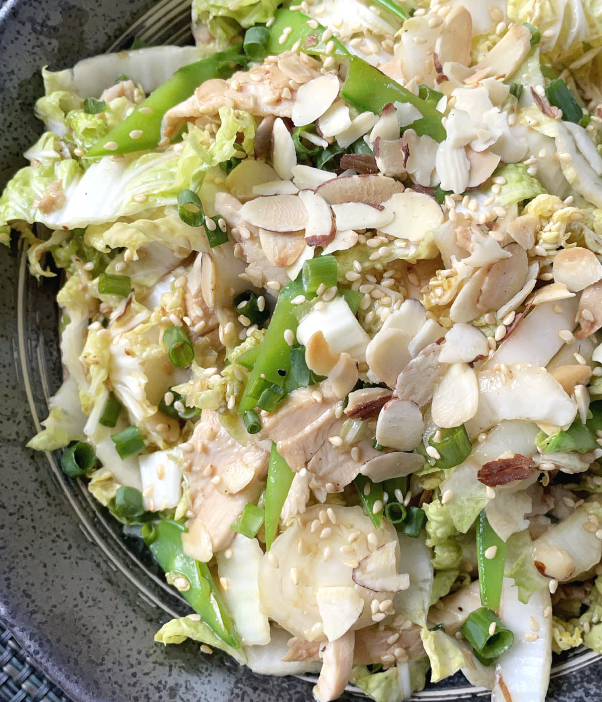 A dark round bowl containing chicken salad topped with sliced almonds, green onions, and sesame seeds.