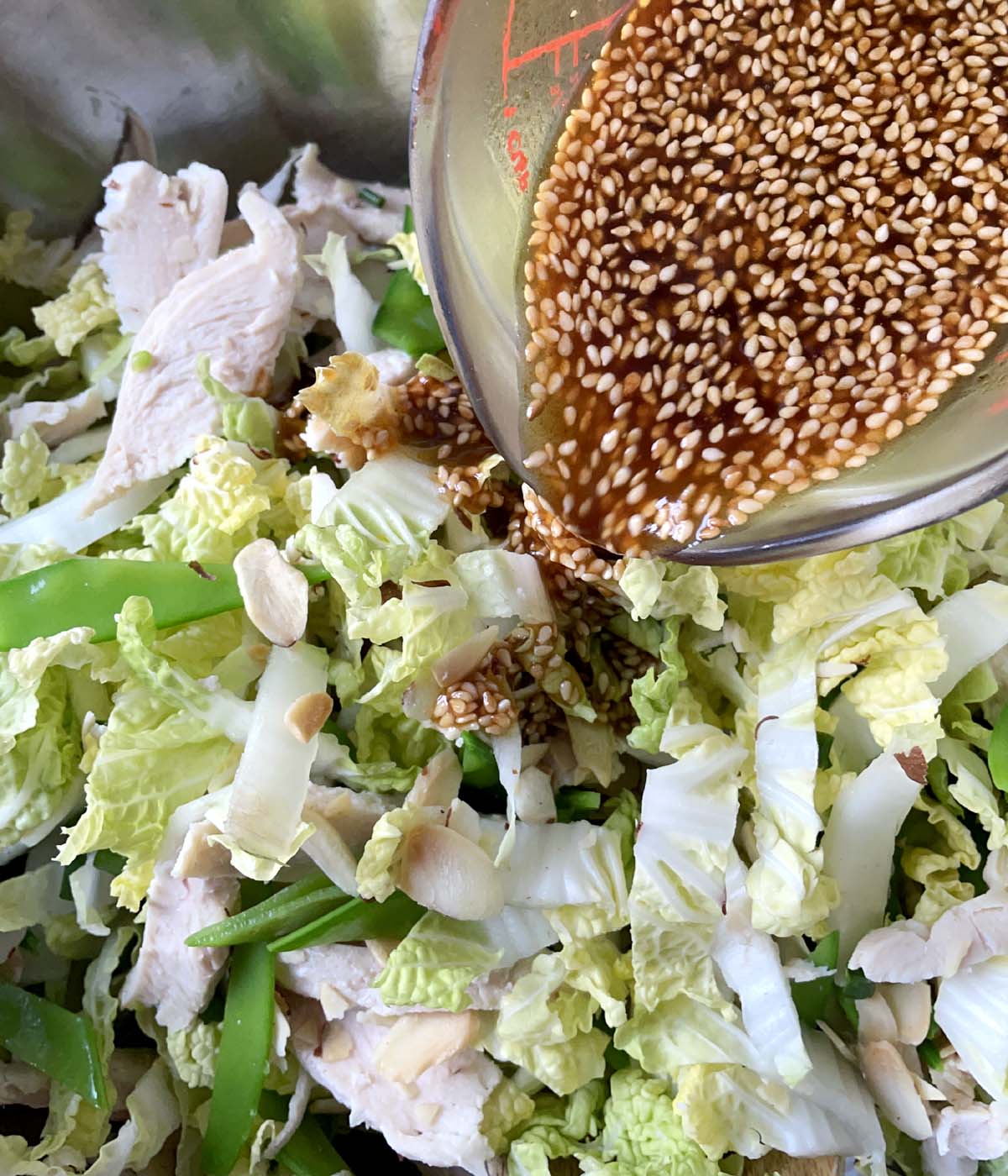 Sesame seed brown dressing being poured into a bowl containing cabbage, sliced almonds, snow peas.