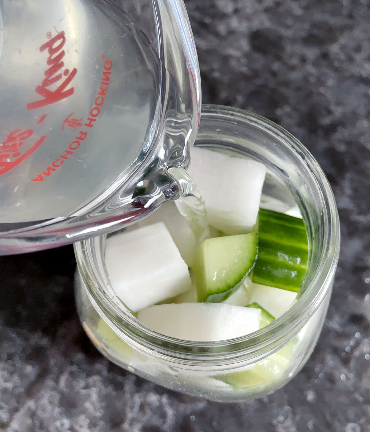 Clear liquid being poured into a jar containing chunks of white daikon radish and green cucumber.