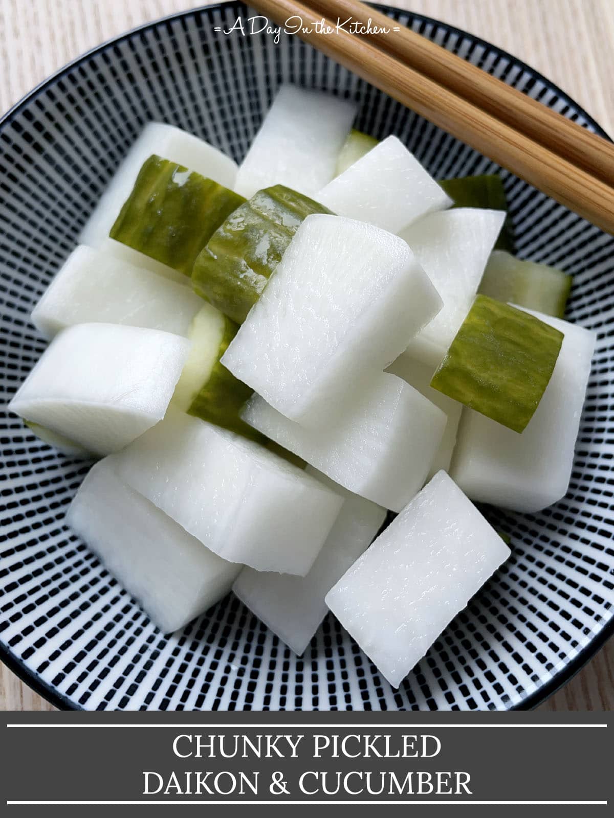 A pair of wooden chopsticks resting on a round bowl containing chunks of white radish and green cucumber, the words chunky pickled daikon and cucumber on the bottom.