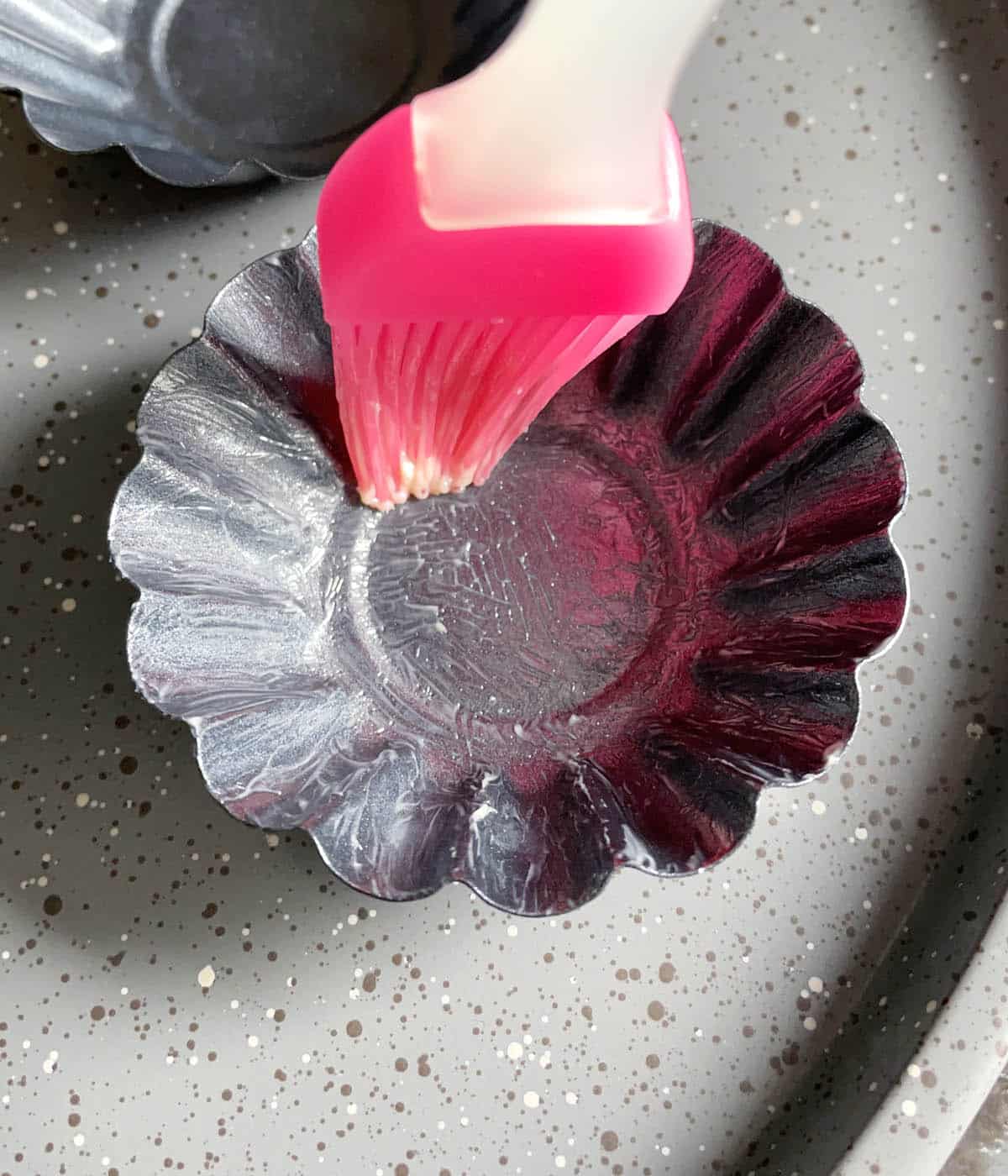 A pink brush spreading butter on a fluted grey dish.