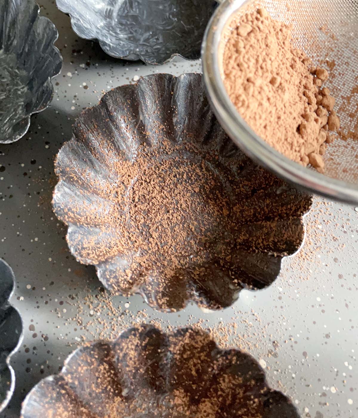 Brown cocoa being sprinkled on grey fluted baking dishes.