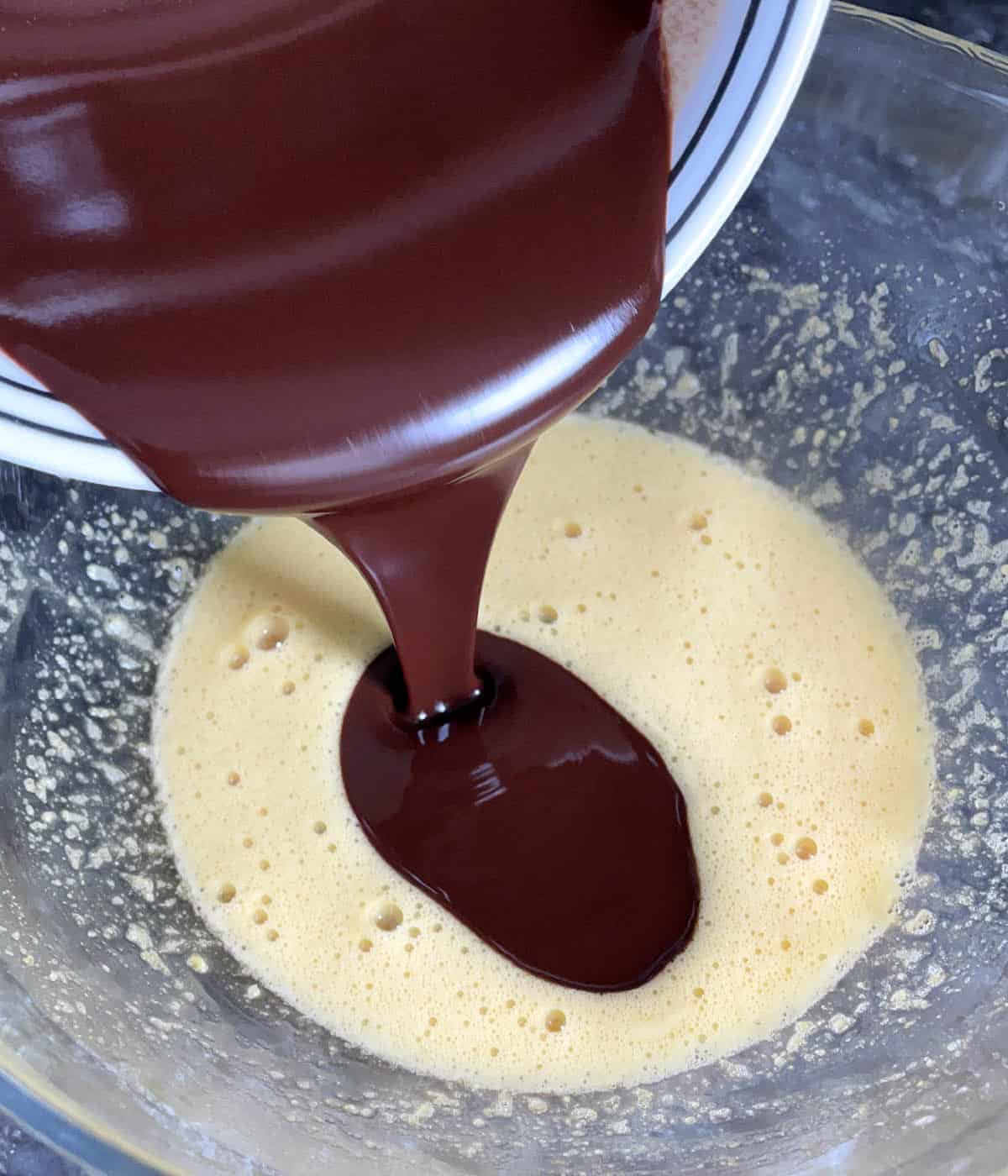 Brown melted chocolate being poured into a glass bowl containing yellow liquid.