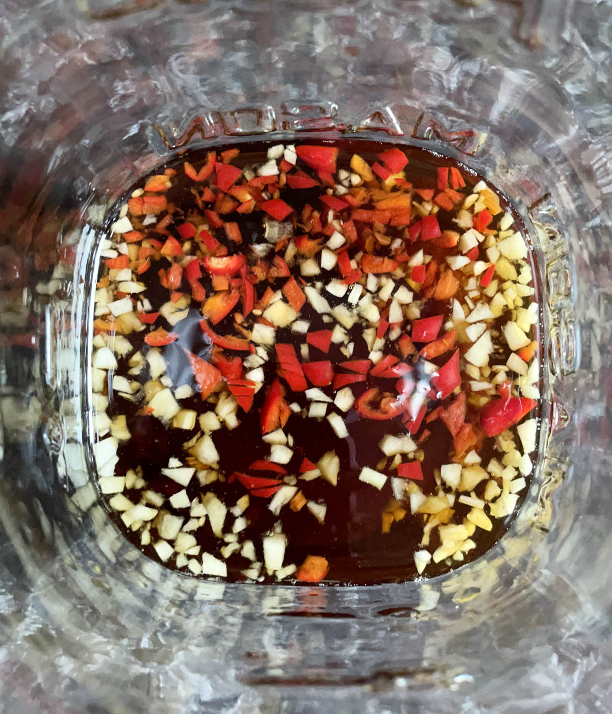 A glass jar containing brown liquid, chopped garlic, and chopped red chili peppers.