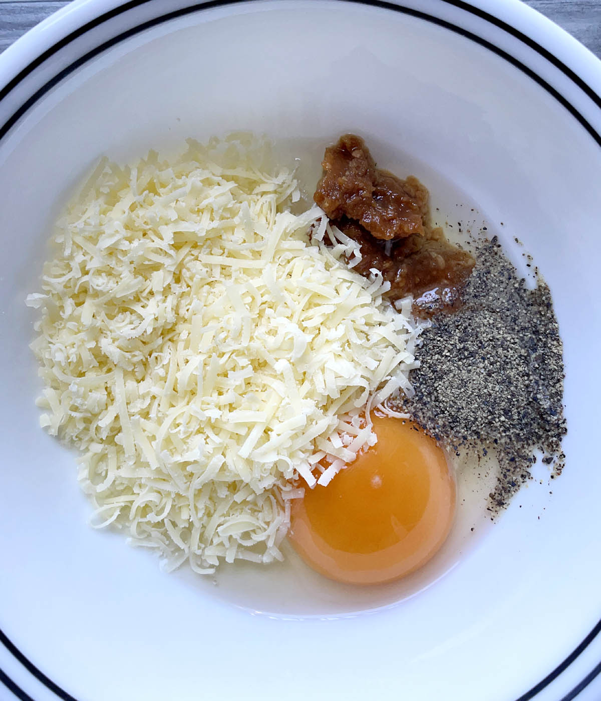 A white bowl containing an egg, black pepper, brown paste, and grated cheese.