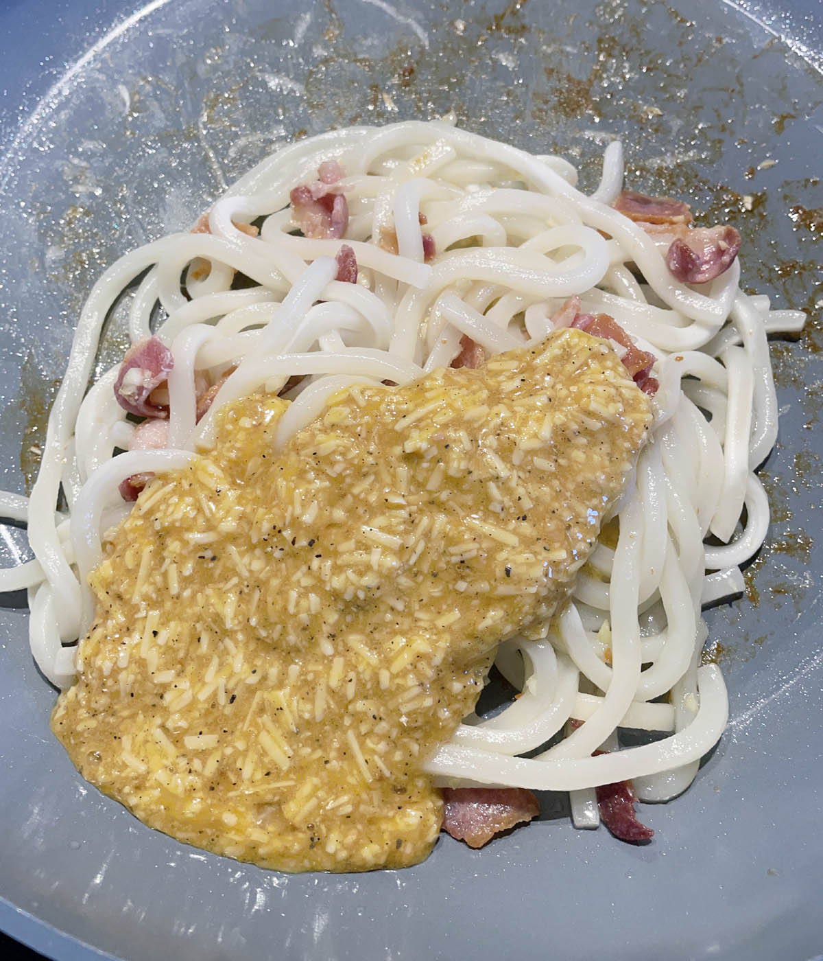 A grey skillet containing cooked bacon pieces, white noodles, and yellow brown sauce.
