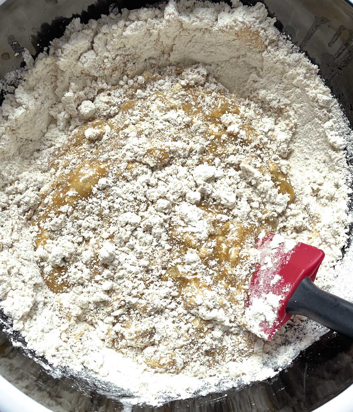 A red rubber spatula mixing white flour and brown liquid in a bowl.
