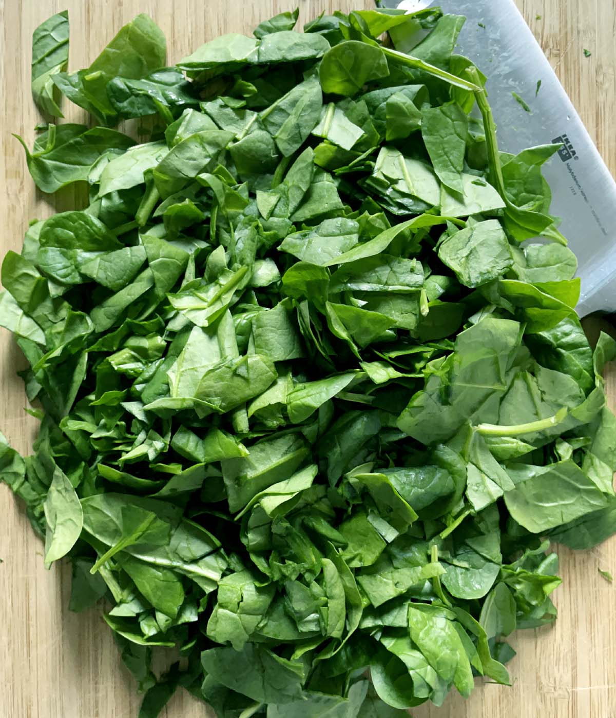 A knife and chopped green spinach on a wooden cutting board.