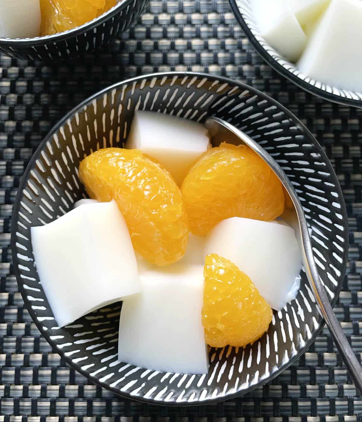A dark round bowl with white cubes and mandarin oranges and a spoon.
