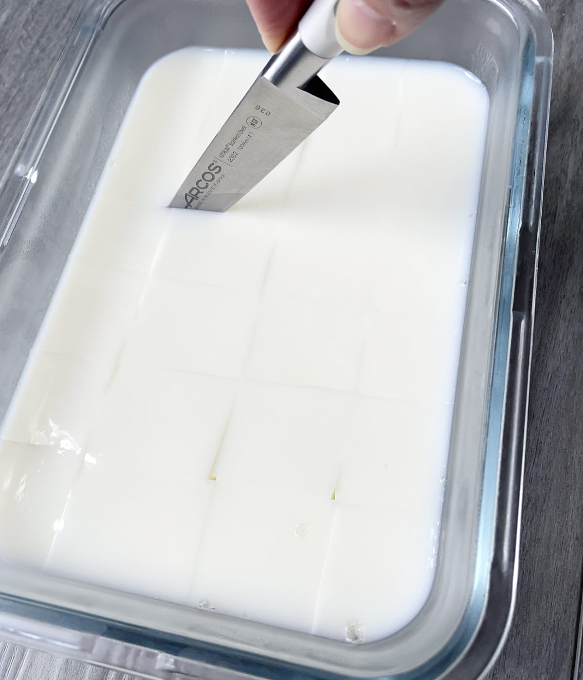 A knife cutting cubes from a glass container of white jelly.