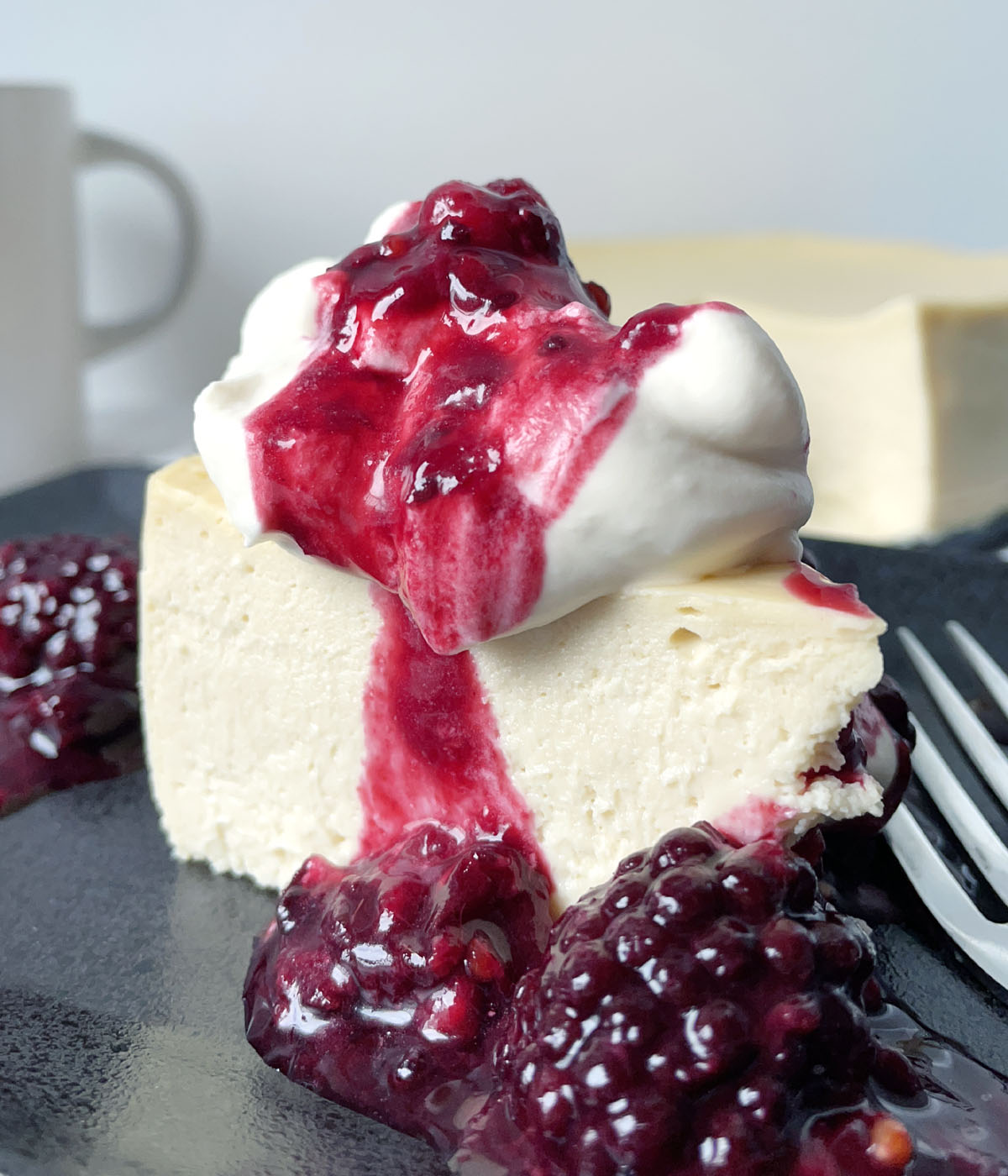 A piece of cheesecake on a dark plate, topped with white whipped cream and red berry sauce.